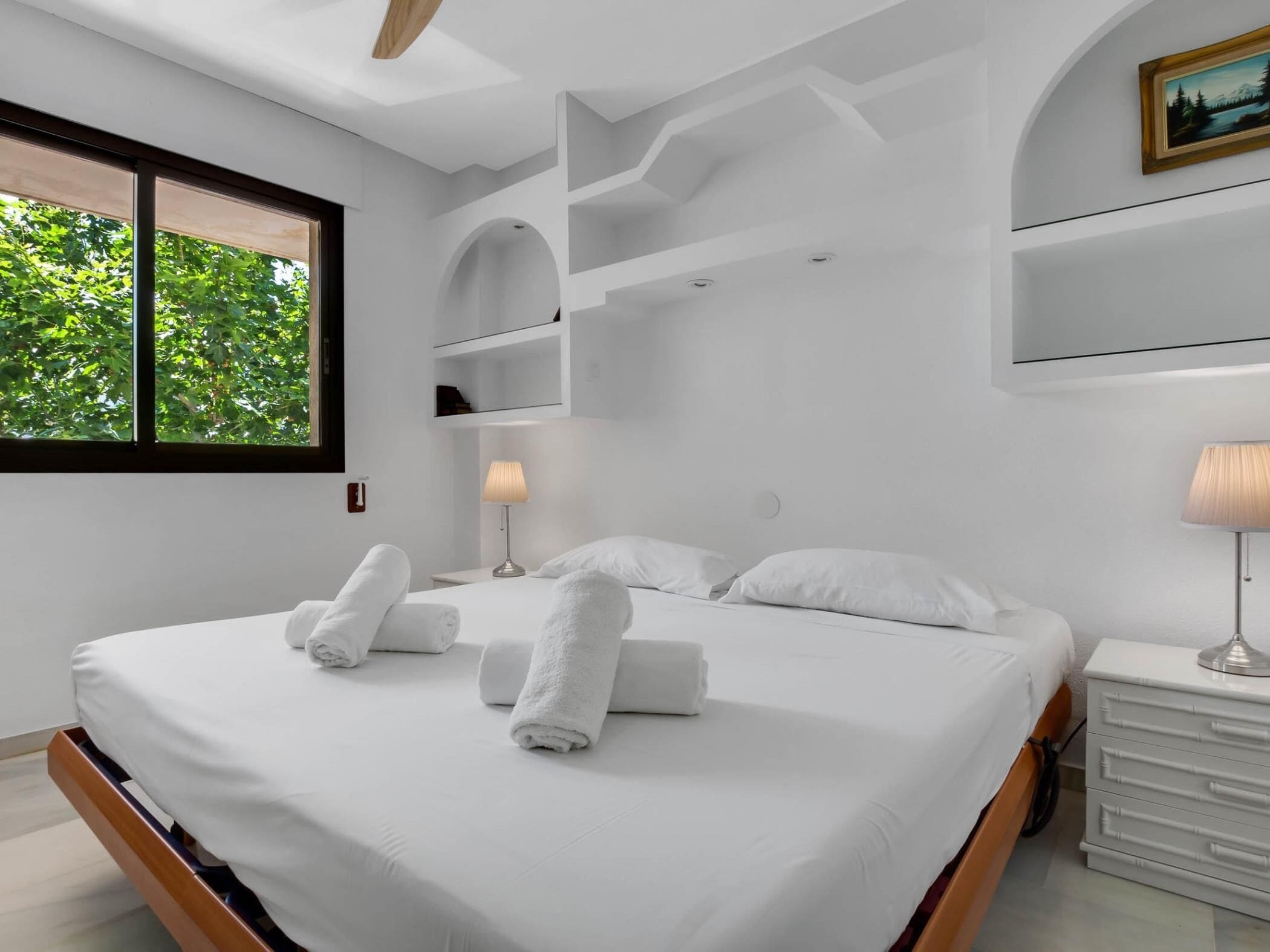 Une chambre moderne avec un grand lit, du linge de lit blanc, une table de chevet avec une lampe et une fenêtre donnant sur un paysage verdoyant.