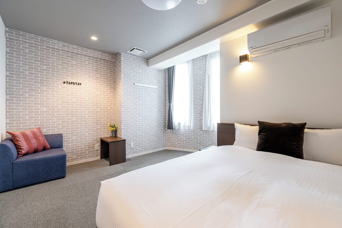 Una habitación de hotel moderna con una cama grande, un sofá azul, una mesita con una planta y una pared de ladrillo.