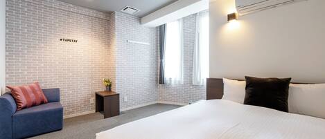 A modern hotel room with a large bed, a blue sofa, a small table with a plant, and a brick wall.