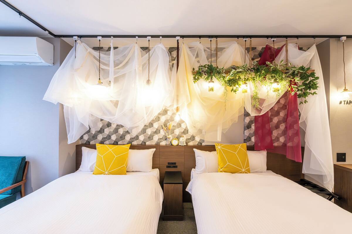 Habitación de hotel con dos camas, cabecera con papel tapiz de estampado geométrico, cortinas translúcidas, plantas colgantes y aire acondicionado de pared.
