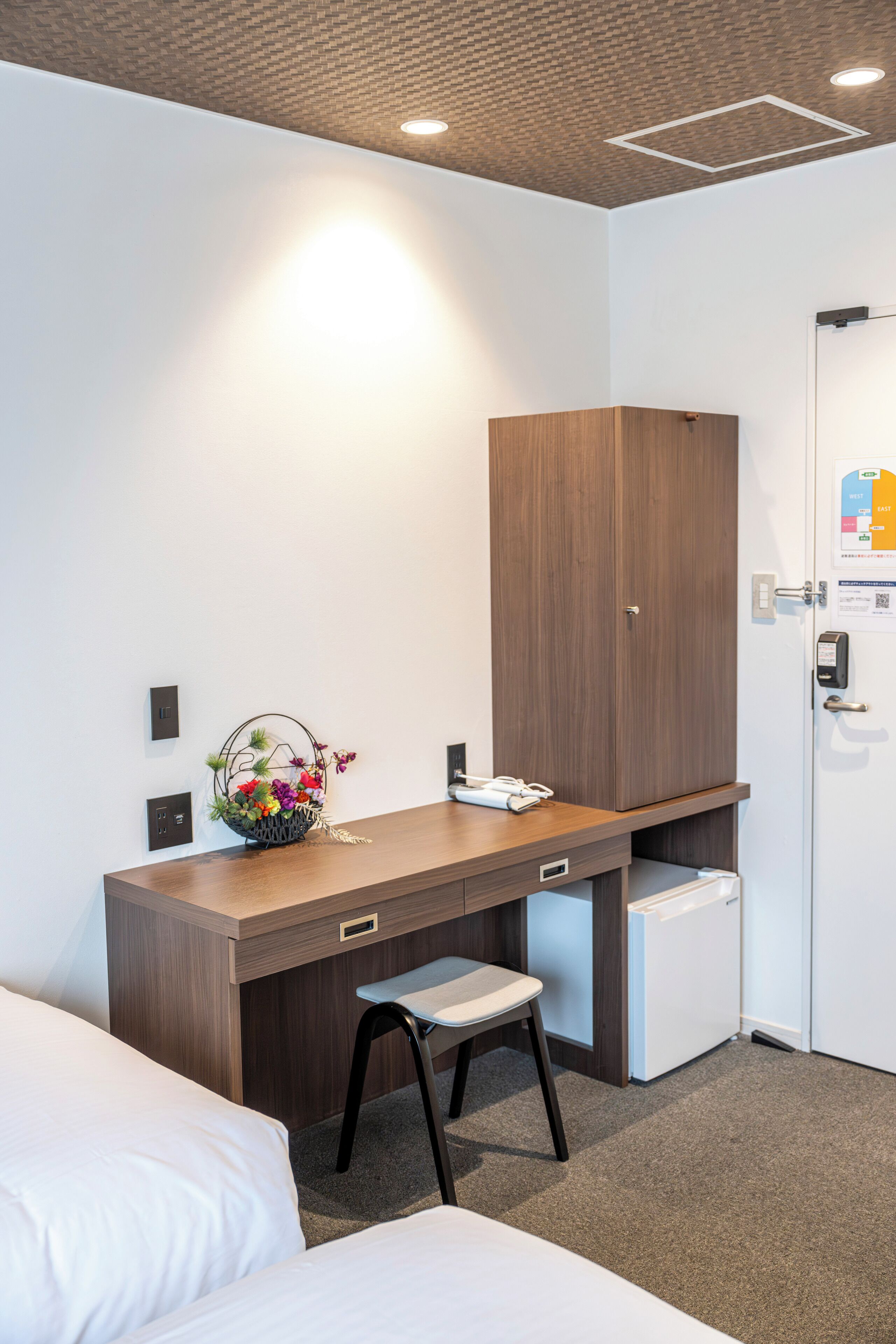 A hotel room with a wooden desk, a small refrigerator, a white bed, and a chair.