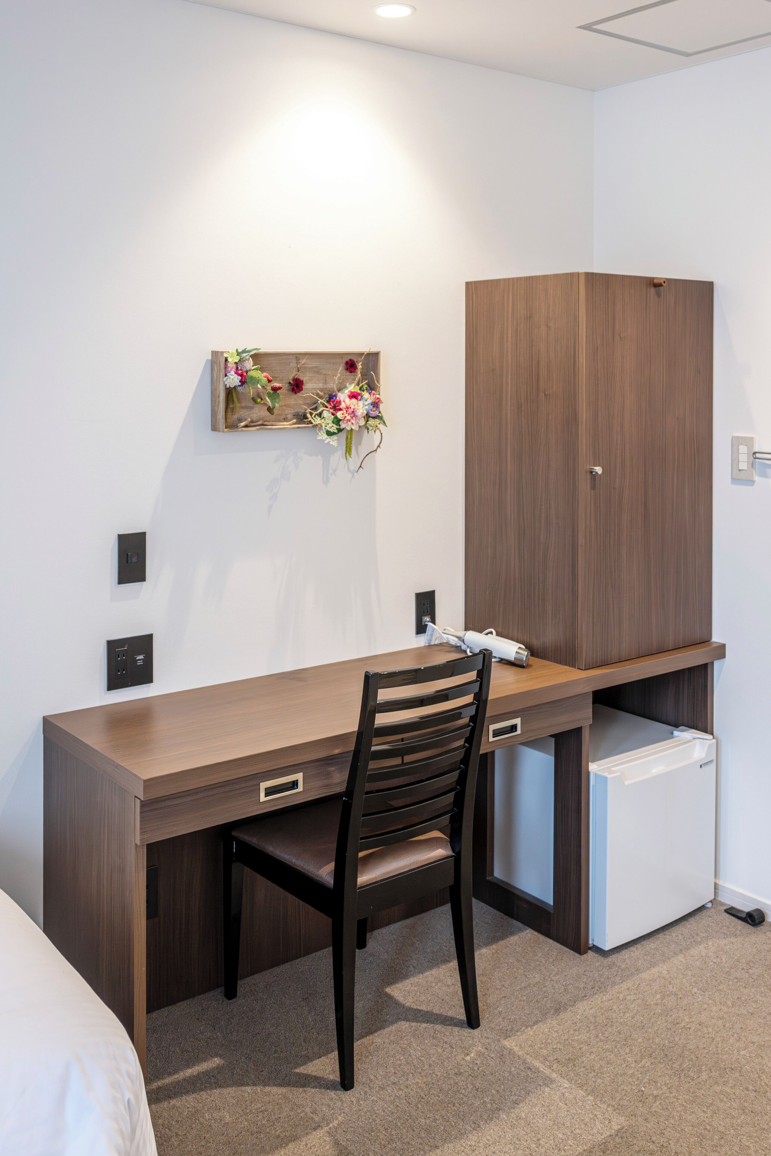 A modern hotel room with a wooden desk, a chair, a small refrigerator, and a wall-mounted artwork.