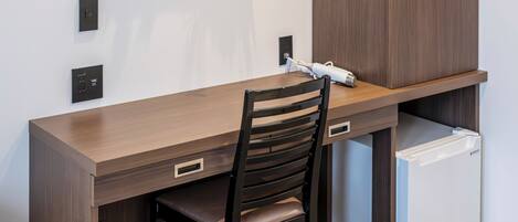 A modern hotel room with a wooden desk, a chair, a small refrigerator, and a wall-mounted artwork.