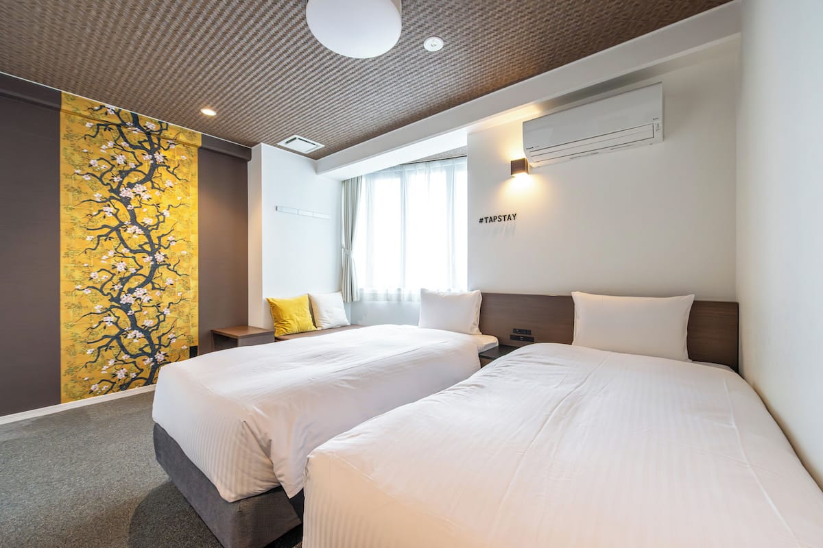 Habitación de hotel con dos camas, un ventanal grande con cortinas, un aire acondicionado de pared y una pared decorativa amarilla con diseño de árbol.