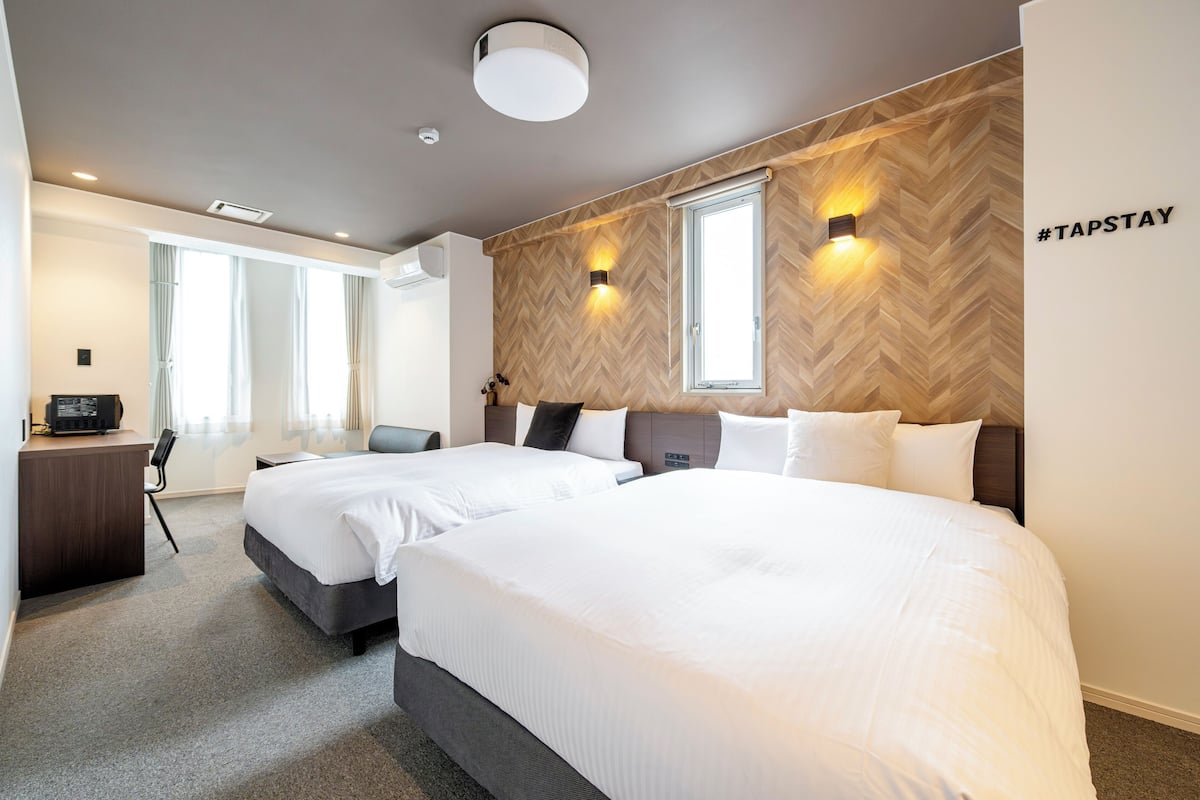 Habitación de hotel con dos camas, un escritorio y una silla. Hay dos ventanas con cortinas, una lámpara de pared y un cartel con el signo de numeral.