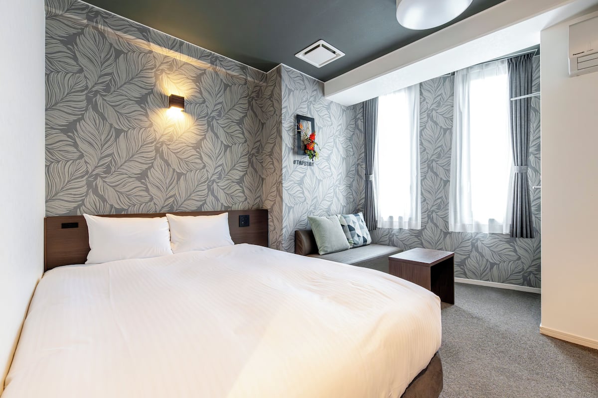 Habitación de hotel moderna con una cama grande, una mesita de noche, un pequeño rincón de descanso y una ventana con cortinas.