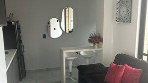 Een moderne woonkamer met een zwarte bank, een witte salontafel, en een zwarte slaapbank met rode kussens. Er staat een witte eettafel met twee witte stoelen, een zwarte koelkast, en een spiegel bevestigd aan de muur.