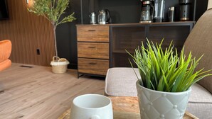 Uma sala de estar moderna com piso de madeira, uma caneca branca em um jogo de bandeja, uma planta verde em um vaso texturizado, um sofá e uma máquina de café em uma prateleira.