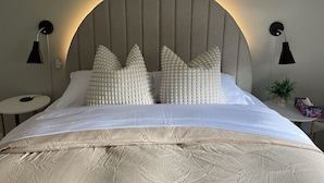 Uma cama bem arrumada com uma cabeceira grande e acolchoada, duas luminárias de cabeceira e um pequeno vaso com planta no criado-mudo.