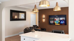 Uma cozinha moderna com ilha central, iluminação pendente, televisão de tela plana na parede e um espaço de estar com sofá e cadeiras.