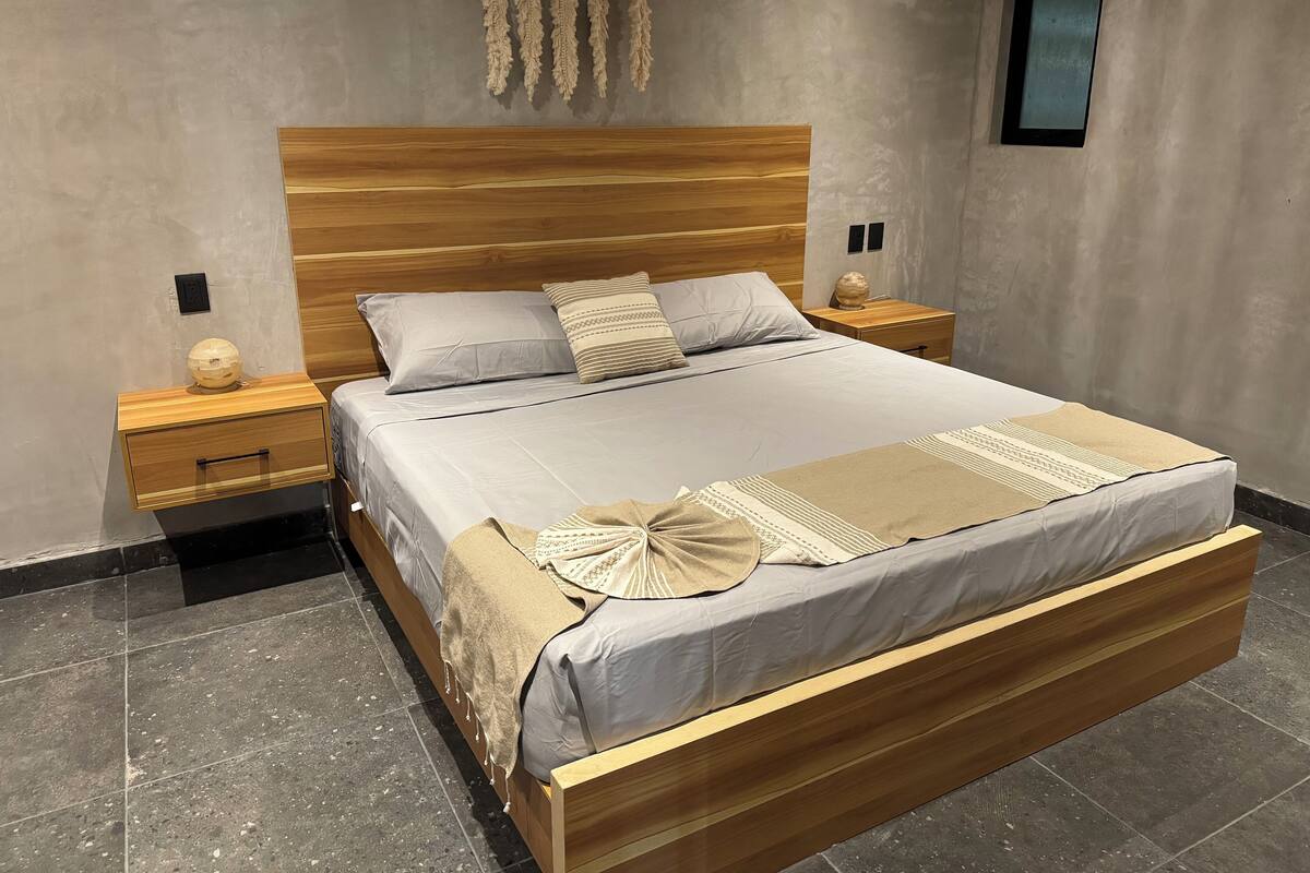 Un dormitorio moderno con cabecera de madera, una cama grande con muchos almohadones, una mesita de noche con lámpara y un adorno en la pared.