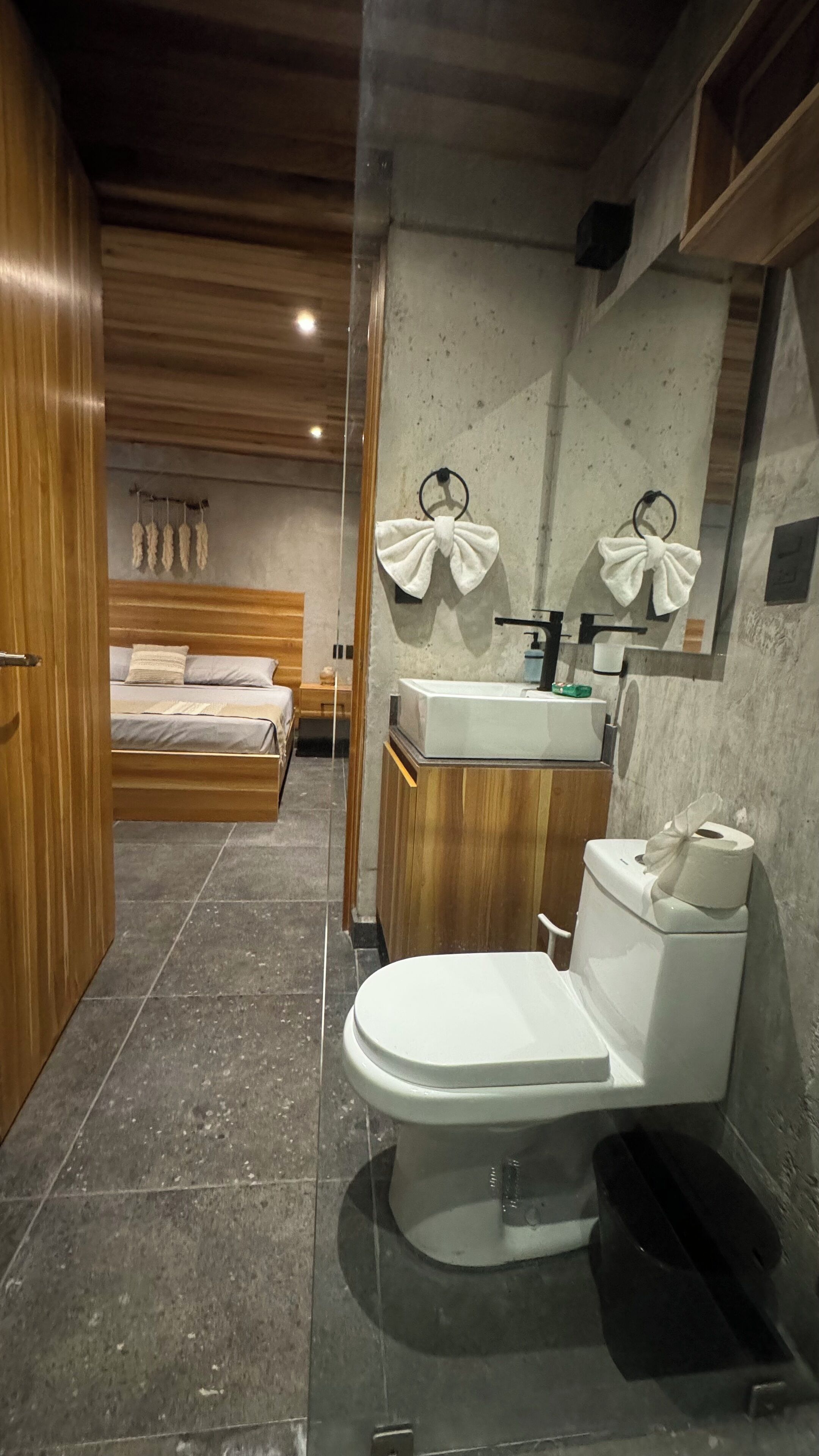 A modern bathroom with a toilet, sink, and mirror. There is a bed visible in the background.