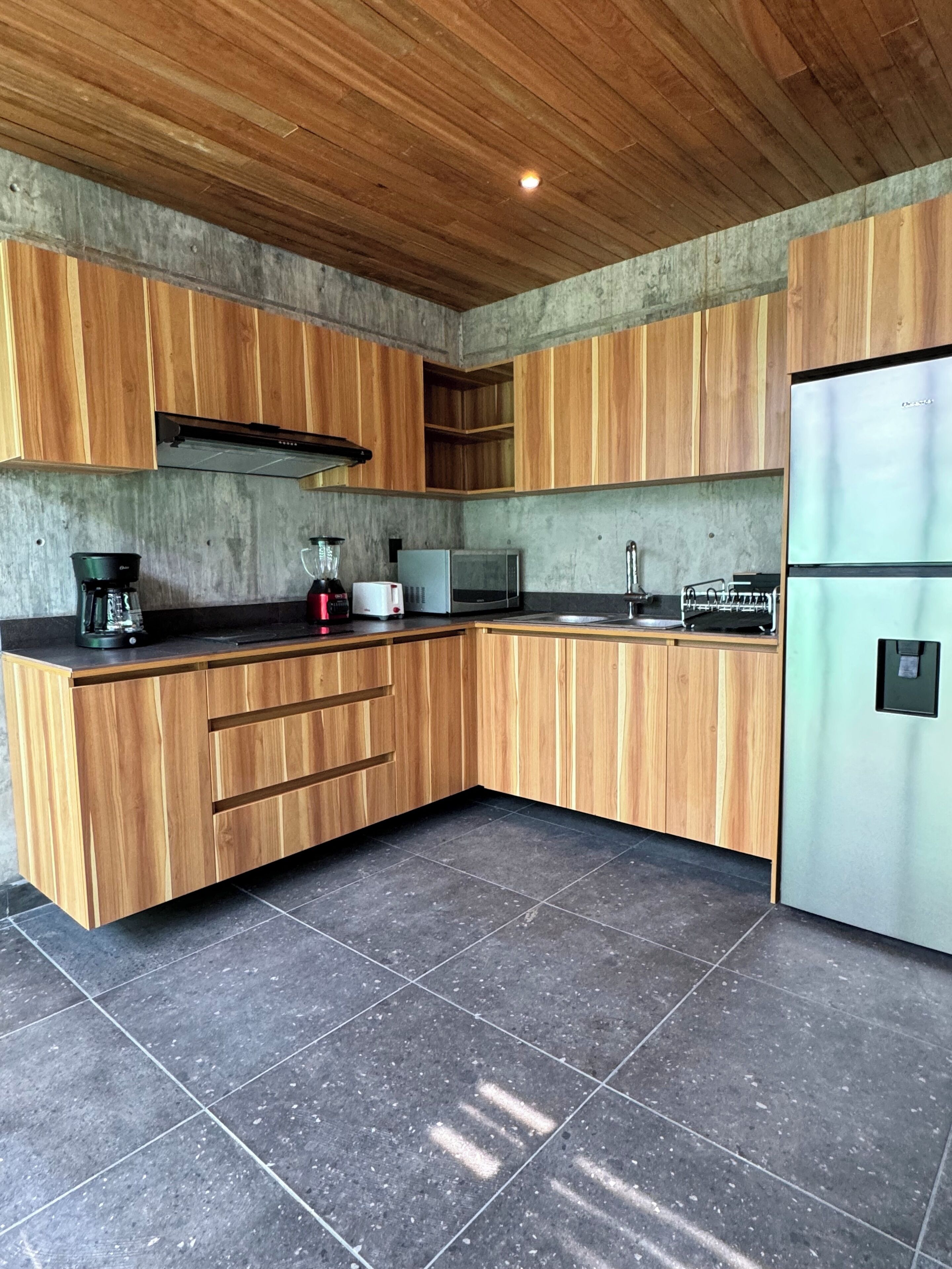 A modern kitchen with wooden cabinets, a stainless steel refrigerator, and a black tiled floor.
