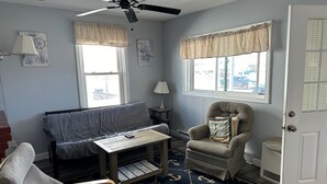 Um quarto com um sofá cinzento, uma mesa de centro de madeira, uma poltrona cinza, um tapete estampado e um ventilador de teto.