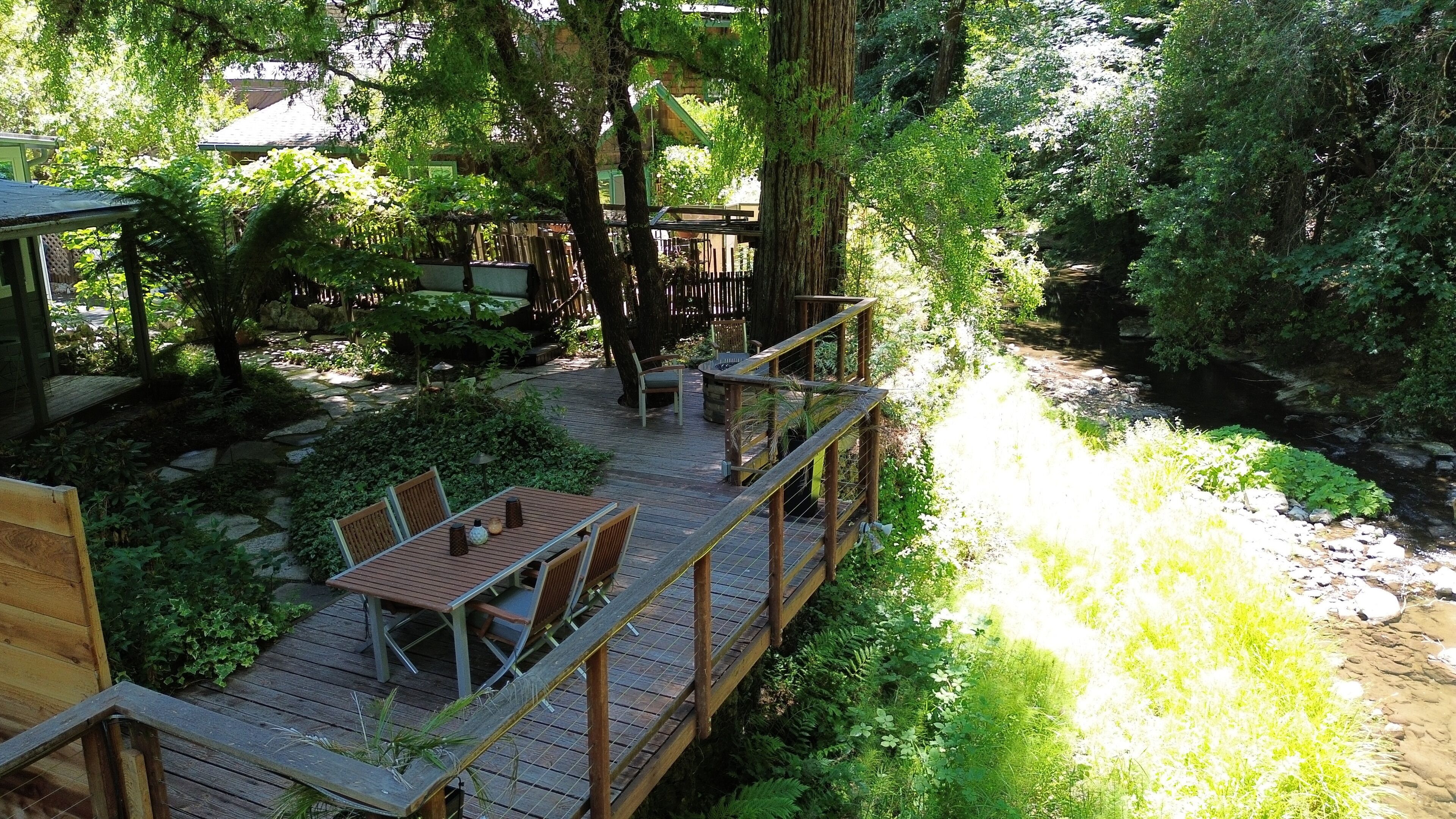Una terraza de madera con una mesa y sillas, rodeada de abundante vegetación y un arroyo.