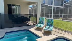 Una piscina con dos sillas de descanso, barandilla de vidrio y vista a una casa al fondo.