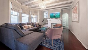 Een woonkamer met een grijze hoekbank, een roze fauteuil, een televisie en een open haard.