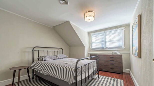 Een slaapkamer met een bed, een dressoir, een klein tafeltje, een raam met jaloezieën en een plafondlamp.