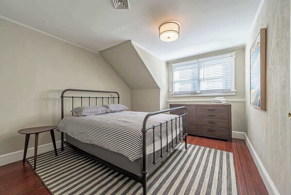 A bedroom with a bed, a dresser, a small table, a window with blinds, and a ceiling light.