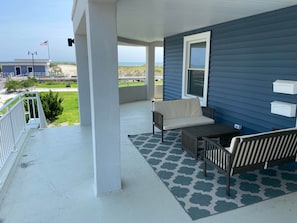 Relax on the covered porch with a view of the beach, featuring a comfortable bench and a small table.