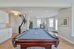 A pool table in a room with a staircase, a dining area, and a large window.