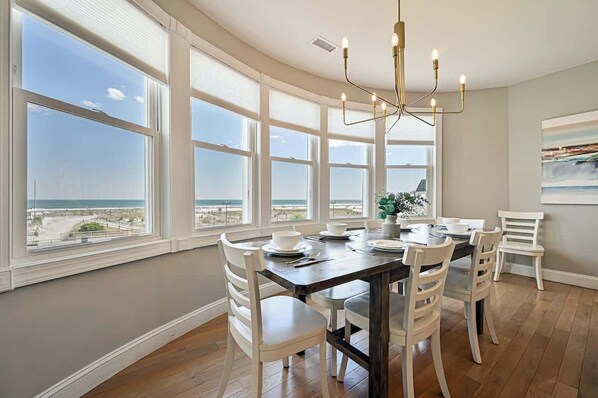 A dining area with a large table, white chairs, and a view of the ocean through large windows. - Unreal ocean Beach Pad - beachfront & boardwalk (Atlantic City)