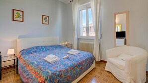 Ein Schlafzimmer mit einem Bett, einem weißen Stuhl, einem Nachttisch, einer Lampe und zwei gerahmten Bildern an der Wand.