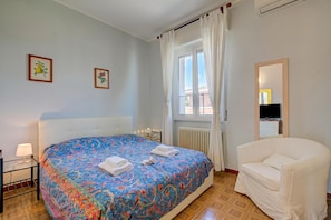 Una habitación de hotel con cama, una silla, ventana con cortinas, radiador y cuadros en la pared.