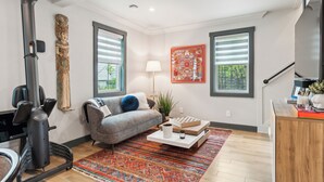 A modern living room with a gray sofa, a colorful rug, a standing fan, and a large window with blinds.