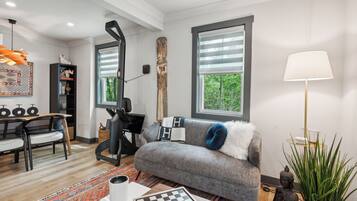 A modern living room with a grey sofa, a coffee table, a chessboard, and a large window with blinds.