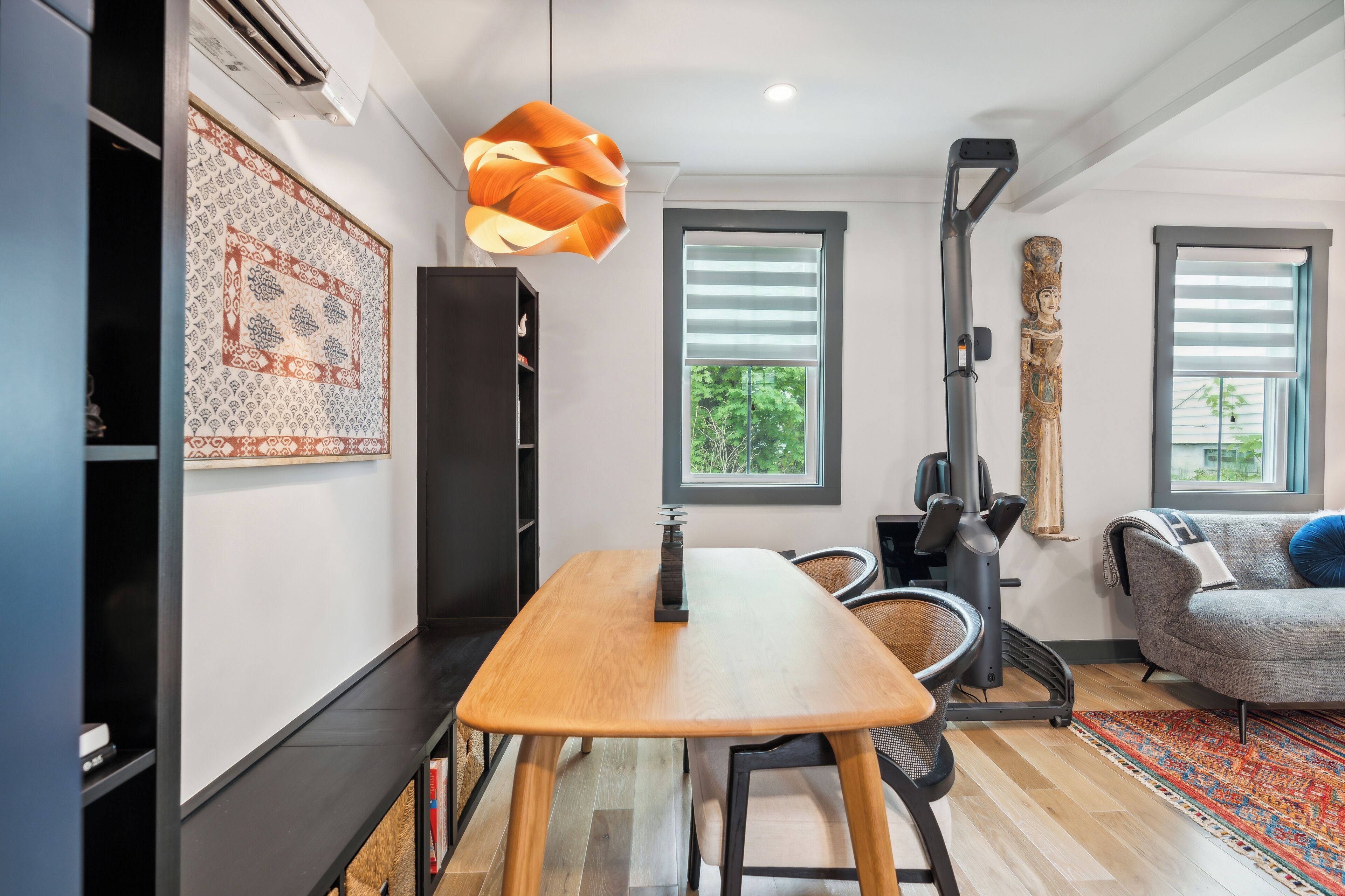 Uma sala de estar moderna com uma mesa de jantar de madeira, um sofá cinza, um tapete estampado e uma grande janela com persianas.