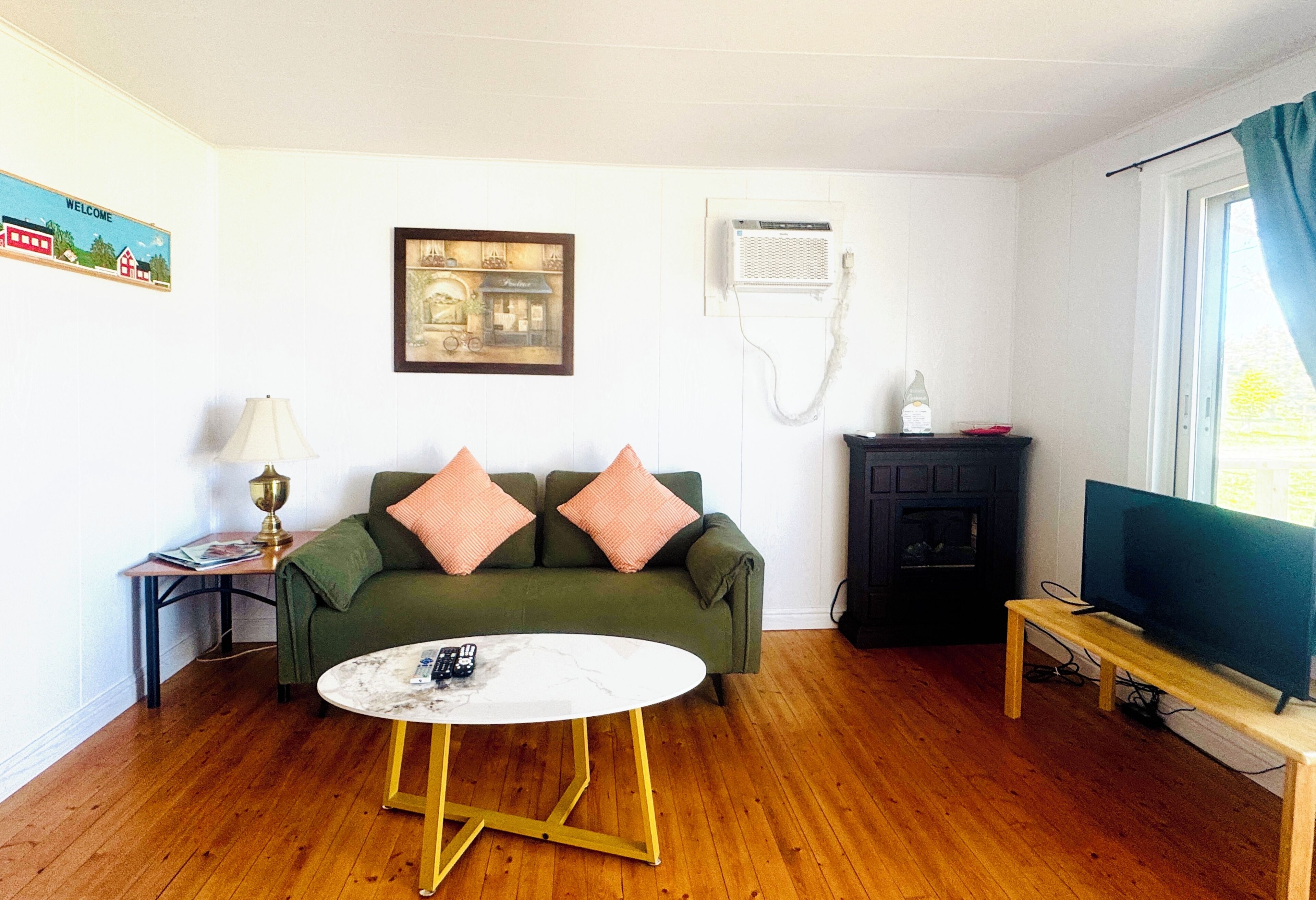 A living room with a green sofa, a white coffee table, a lamp, a fireplace, a television on a stand, and a wall-mounted air conditioner.