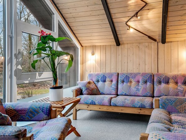 Un interior moderno con techo de madera, sofá estampado, planta en maceta y vistas a través de ventanas amplias.