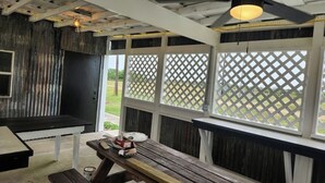 Ett takförsett utomhusområde med sittplatser, en träbänk och ett picknickbord, en takfläkt och utsikt genom ett fönster med ett dekorativt gallerverk.