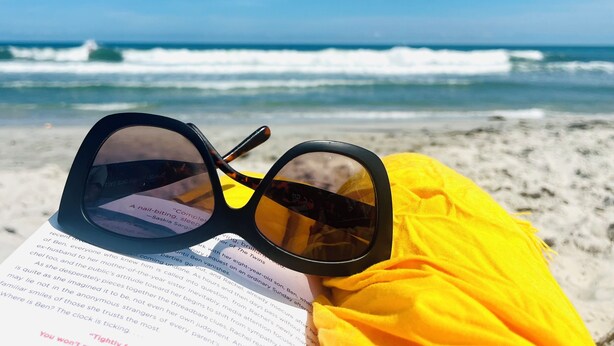 Une paire de lunettes de soleil et un livre sur une plage, avec les vagues de l’océan en arrière-plan.
