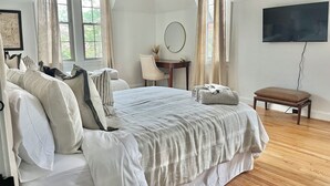 Ein Schlafzimmer mit einem großen Bett, einem Schreibtisch, einem Sessel, einem Fernseher und einem Fenster mit Vorhängen.