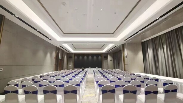 Uma sala de conferências com fileiras de cadeiras brancas, cada uma com uma fita azul. A sala possui um teto moderno com iluminação embutida e um fundo escuro na frente.