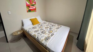 Un lit simple avec une couvre-lit à motifs floraux, un oreiller blanc et un oreiller de couleur moutarde. Il y a une petite table ronde sur laquelle se trouve un téléphone. Un tableau abstrait aux couleurs vives est accroché au mur, au-dessus du lit.