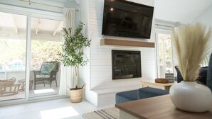 Una sala de estar con televisor montado en la pared, chimenea, ventana grande y planta en maceta.