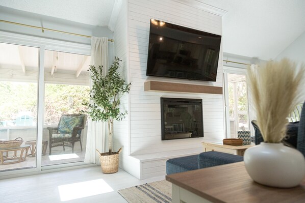 A living room with a TV mounted on the wall, a fireplace, a large window, and a potted plant.