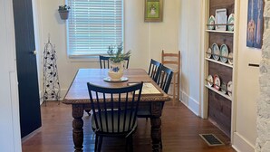 A dining room with a wooden table, chairs, a chandelier, a window with blinds, a plant, and a shelf with decorative items.
