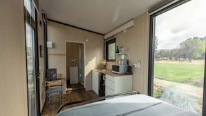 Een kleine kamer met een bed, een kitchenette en een groot raam met uitzicht op een gazon.