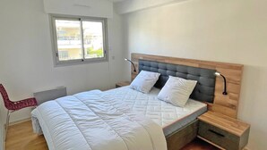 Un dormitorio con cama, mesita de noche, silla, ventana y lámpara de pared.