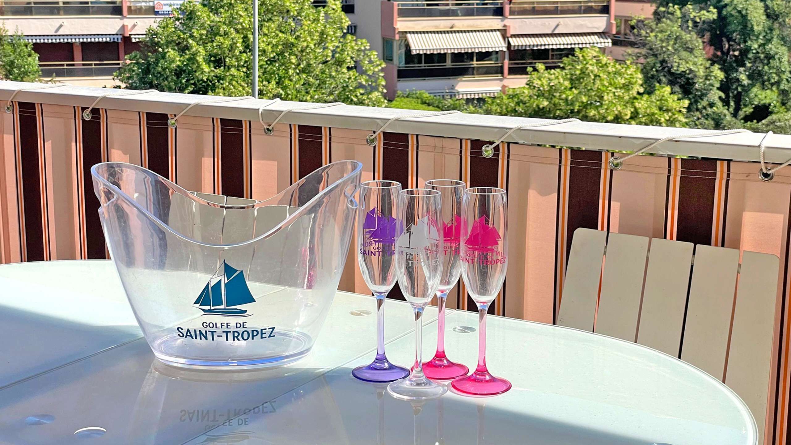A table with a clear pitcher and three wine glasses on a balcony with a view of buildings and trees.