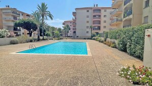 Una piscina rodeada por un patio cubierto de guijarros, con palmeras y un edificio de varios pisos al fondo.