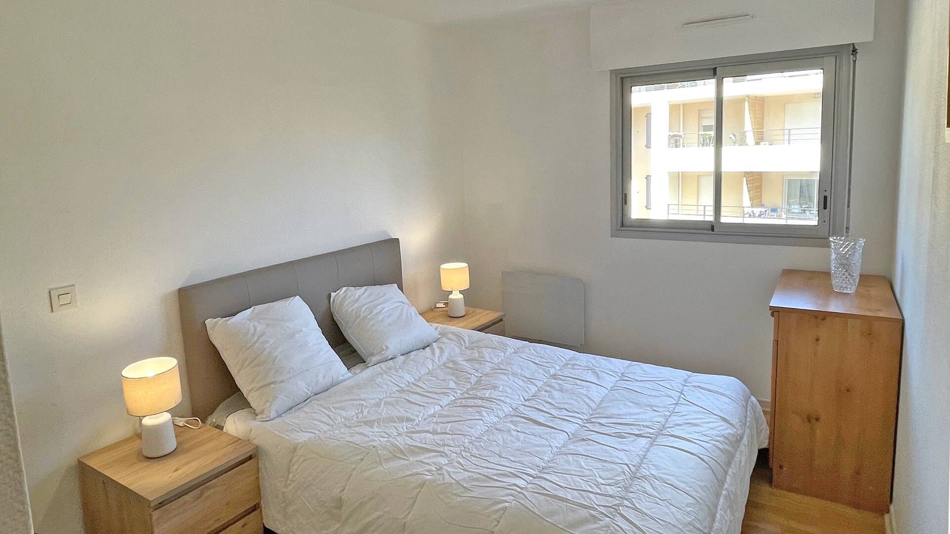 Una cama bien tendida con sábanas blancas en una habitación con ventana y mesita de noche de madera con lámpara.
