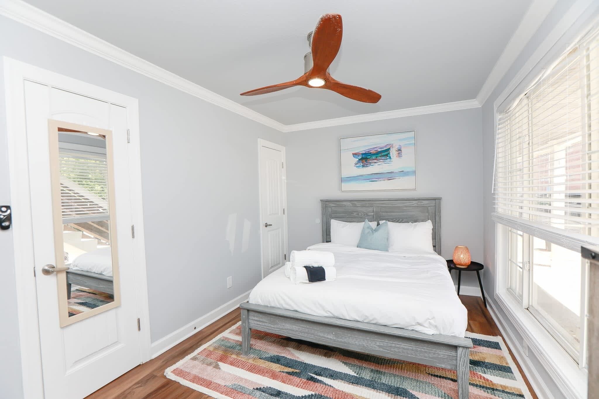 Un dormitorio con cama, ventilador de techo, ventana con persianas, puerta y un cuadro en la pared.