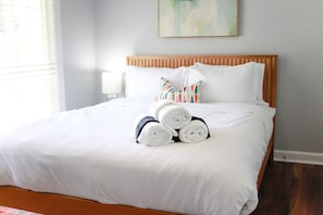 A neatly made bed with white linens and rolled towels, a wooden headboard, and a single pillow with a colorful pattern.