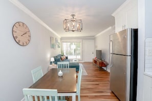 A modern kitchen with a large refrigerator, a dining table with chairs, and a clock on the wall. - Sojourn Isle Suite 2 BR 2 BA at Beach Cottages (Norfolk)