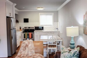 A compact kitchen with white cabinets, a stainless steel refrigerator, a microwave, and a stove. There is a dining area with a white table and chairs, and a brown sofa with a fluffy throw blanket. - Sojourn Palm Suite at Beach Cottages (Norfolk)